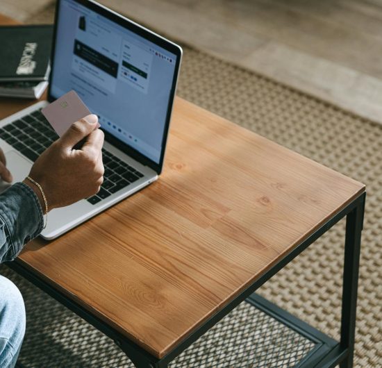 Person holding a credit card while shopping online using a laptop on a wooden table.