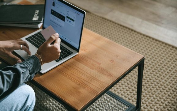 Person holding a credit card while shopping online using a laptop on a wooden table.