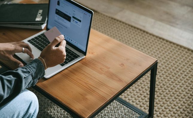 Person holding a credit card while shopping online using a laptop on a wooden table.